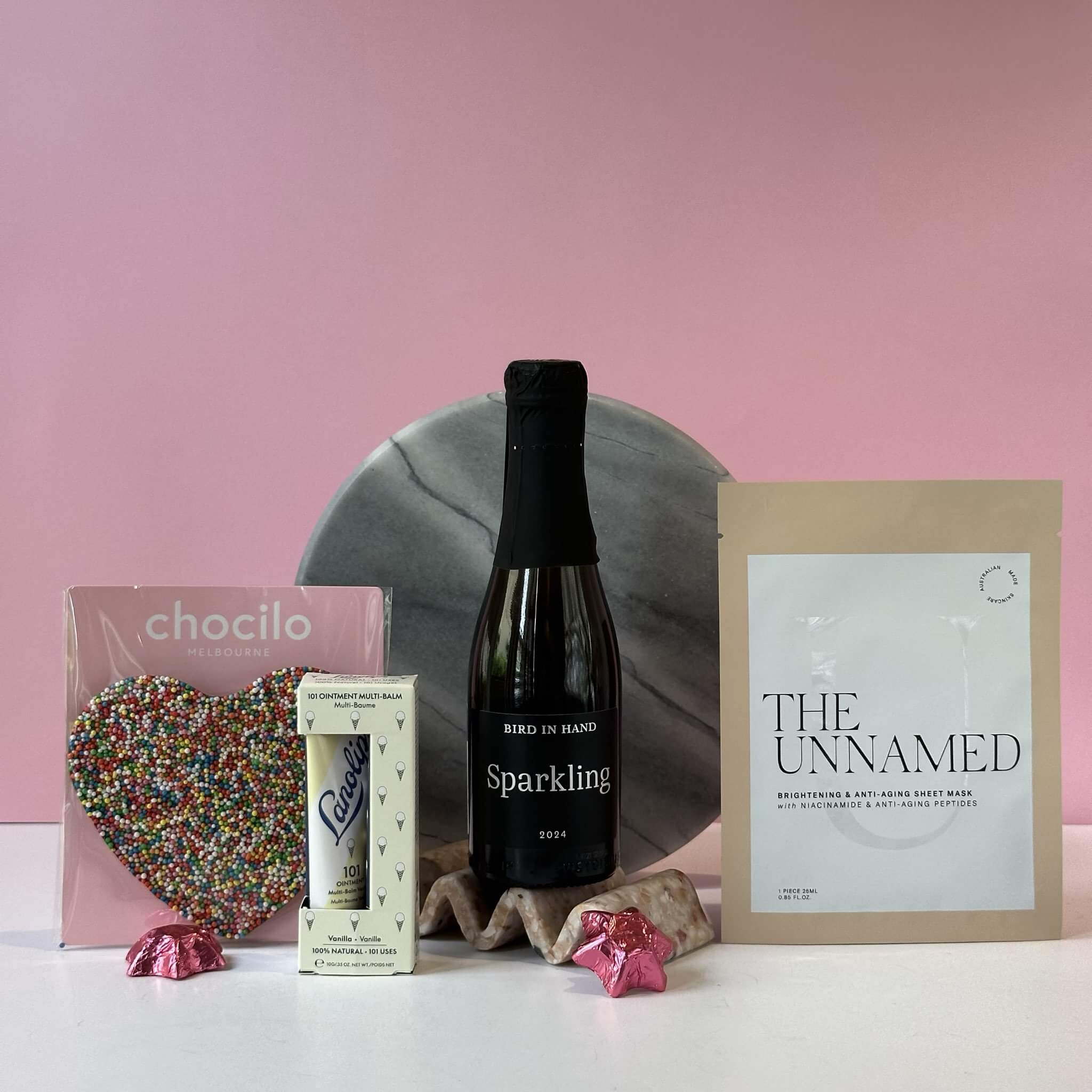 The Best Friend gift bundle displayed against a pink background, featuring a Chocilo Melbourne chocolate heart with colorful sprinkles, a Bird in Hand Sparkling wine bottle (2024), a Lanolips vanilla lip balm, and The Unnamed brightening sheet mask, arranged on a marble surface with decorative pink foil chocolates.